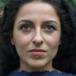 Op de afbeelding: Een close-up portret van een persoon met donker haar en bruine ogen. Ze draagt een donkerblauw shirt en heeft een klein gouden oorbelpiercing in haar onderlip.