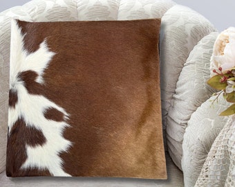 May include: A brown and white cowhide throw pillow with a soft, plush texture. The pillow is shown on a white couch with a floral pattern.