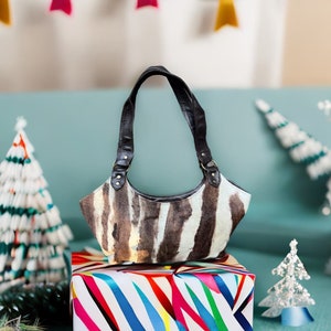 May include: A small, brown and white zebra-print handbag with black leather straps and gold hardware. The bag is sitting on a colorful striped gift box.