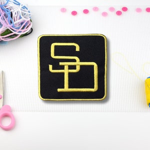 May include: Black embroidered patch with gold lettering that reads "SD".