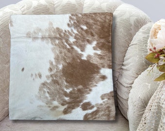 May include: A brown and white cowhide pillow with a soft, plush texture. The pillow is sitting on a white, tufted armchair.