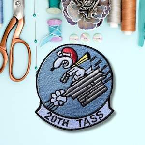 May include: A blue and white embroidered patch with a cartoon dog wearing a red hat and holding a gun. The dog is flying in a plane with the text "20th TASS" on the bottom.