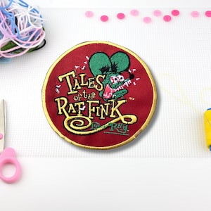 May include: A round, red embroidered patch with the text "Tales of the Rat Fink" and a green cartoon rat. The patch has a gold border. Pink scissors, buttons, and yarn are in the background.