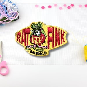 May include: A yellow and red embroidered patch with the words "Rat Fink" and a cartoon rat with green eyes and a red mouth. The rat is holding a wrench and has a pink body. The patch has a black border and the words "Rat Fink" are in a black font.