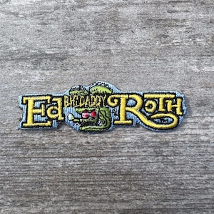 May include: An embroidered patch featuring the name "Eddie Roth" in yellow lettering with a green cartoon monster in the center. The monster has red eyes and is wearing a blue hat.