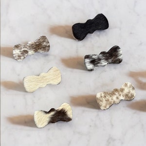 May include: Six different cowhide bow tie hair clips. The hair clips are in various shades of brown, black, and white.