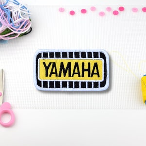 May include: A black and white embroidered patch with a yellow background. The patch says "YAMAHA" in black lettering.