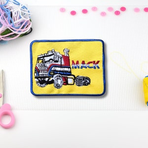 May include: A yellow embroidered patch with a blue border featuring a red, white, and blue semi-truck with the word "MACK" in blue lettering.