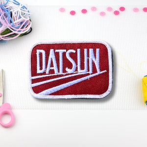 May include: Red embroidered patch with white stitching. The patch features the word "DATSUN" in white lettering.