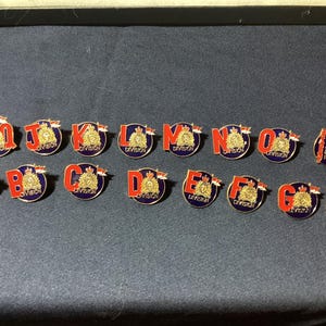 May include: A collection of round, enamel pins with a gold emblem and red lettering. Each pin features a letter from A to O, with the letters set against a blue background. The pins are likely collectible items.