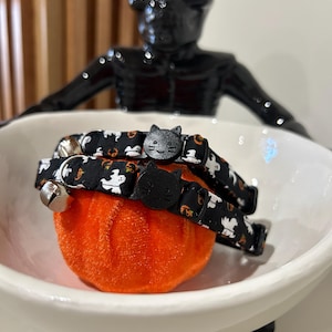 May include: Two black cat collars with Halloween patterns, including ghosts and pumpkins, are displayed in a white bowl. Each collar has a black cat-shaped buckle and a silver bell. The collars rest on a small orange pumpkin.