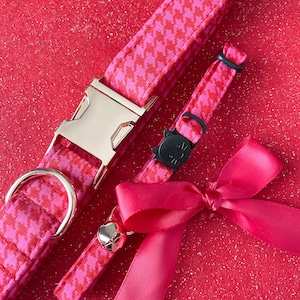 K&ouml;nnte beinhalten: Ein pinkfarbenes und rotes Hahnentrittmuster-Halsband f&uuml;r Katzen mit einer goldenen Schnalle und ein passendes pinkfarbenes und rotes Hahnentrittmuster-Abnehmbares Halsband f&uuml;r Katzen mit einem schwarzen Katzenanh&auml;nger. Ein rosafarbenes Satinband mit Schleife ist im Lieferumfang enthalten.