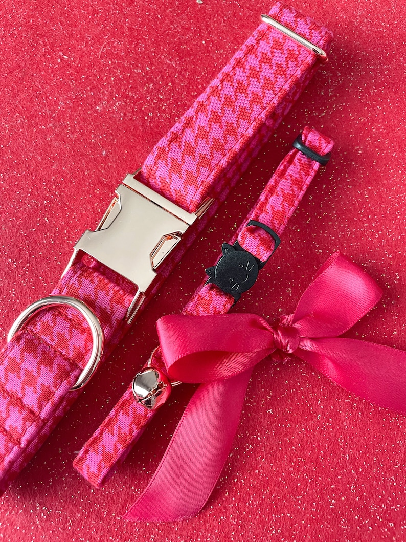 K&ouml;nnte beinhalten: Ein pinkfarbenes und rotes Hahnentrittmuster-Halsband f&uuml;r Katzen mit einer goldenen Schnalle und ein passendes pinkfarbenes und rotes Hahnentrittmuster-Abnehmbares Halsband f&uuml;r Katzen mit einem schwarzen Katzenanh&auml;nger. Ein rosafarbenes Satinband mit Schleife ist im Lieferumfang enthalten.