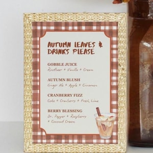 May include: A framed sign with a gold border and a brown and white checkered pattern. The sign reads "Autumn Leaves Drinks Please" and lists four drink recipes: Gobble Juice, Autumn Blush, Cranberry Fizz, and Berry Blessing. A drawing of a drink is in the bottom right corner.