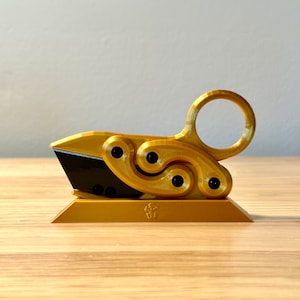 May include: A gold-colored metal paper cutter with a unique, intricate design. The cutter has a ring handle and a sharp blade. It is sitting on a gold-colored base with a small symbol on it.