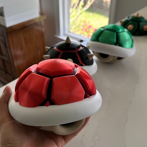 May include: Three 3D printed Super Mario Bros. Koopa Shell power-ups. The shells are red, green, and black with white undersides.