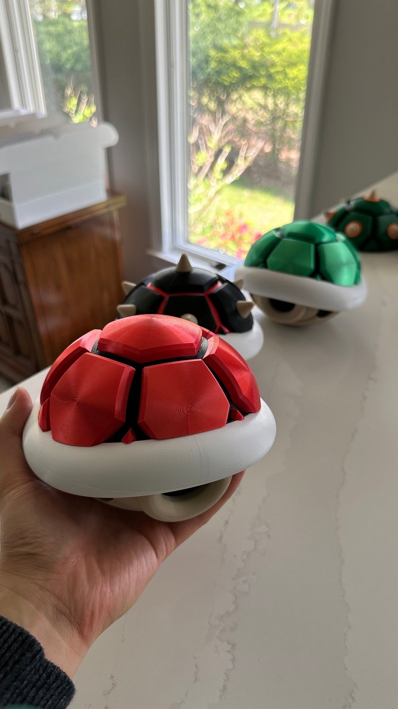 May include: Three 3D printed Super Mario Bros. Koopa Shell power-ups. The shells are red, green, and black with white undersides.