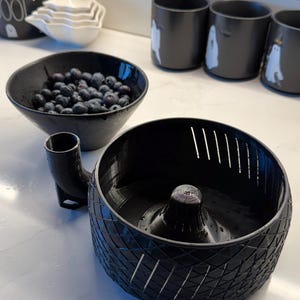 May include: A black berry washing and draining bowl with a unique design, alongside a bowl of blueberries. The bowl has a built-in spout and a textured exterior. Several black ghost-themed mugs are in the background.