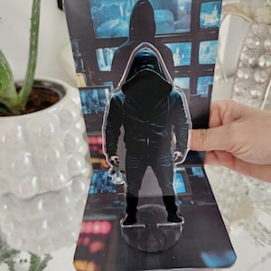Könnte beinhalten: Ein transparenter Acryl-Aufsteller mit einer Figur in schwarzer Kapuzenjacke und dunkler Jeans, die vor einem Hintergrund steht, der einen Raum mit mehreren Computermonitoren zeigt, auf denen Überwachungsaufnahmen zu sehen sind.