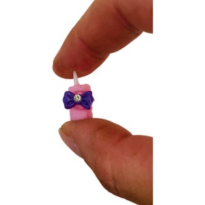 Könnte beinhalten: Eine kleine rosa Kerze mit einer lila Schleife und einem Strassstein in der Mitte. Die Kerze hat einen weißen Docht und wird zwischen zwei Fingern gehalten.