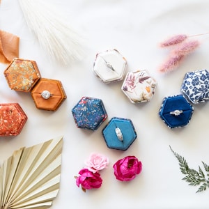 May include: A collection of hexagonal ring boxes in various colors and patterns, including solid orange, blue, and white velvet, as well as floral prints. Several silver rings are displayed inside the open boxes, arranged on a white surface.