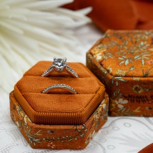 May include: A hexagonal ring box with a floral pattern in shades of brown, orange, and green. The box is open, revealing a velvet interior with two rings. One ring is a solitaire diamond engagement ring, and the other is a simple band.