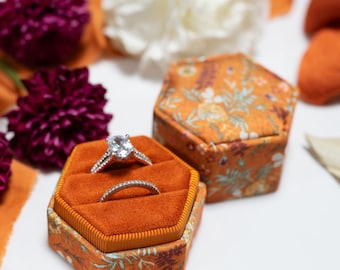 Könnte beinhalten: Eine sechseckige Ringbox mit einem samtigen Innenfutter in gebranntem Orange und einem floral gemusterten Äußeren, die zwei silberfarbene Ringe mit klaren Steinen enthält. Die Box ist von dunkelvioletten Chrysanthemen und orangefarbenem Stoff umgeben.