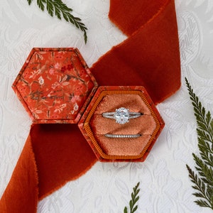 May include: A hexagonal ring box with a floral pattern on the outside and a peach velvet interior. The box is open and contains a silver engagement ring and a wedding band.