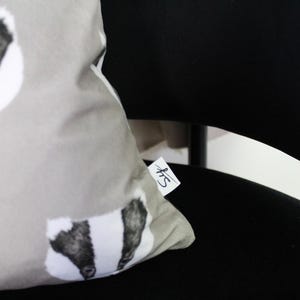 Puede incluir: Un coj&iacute;n decorativo gris claro con un dise&ntilde;o de tej&oacute;n. El tej&oacute;n tiene marcas en blanco y negro. El coj&iacute;n est&aacute; sobre una silla negra. Se adjunta una peque&ntilde;a etiqueta con texto manuscrito.