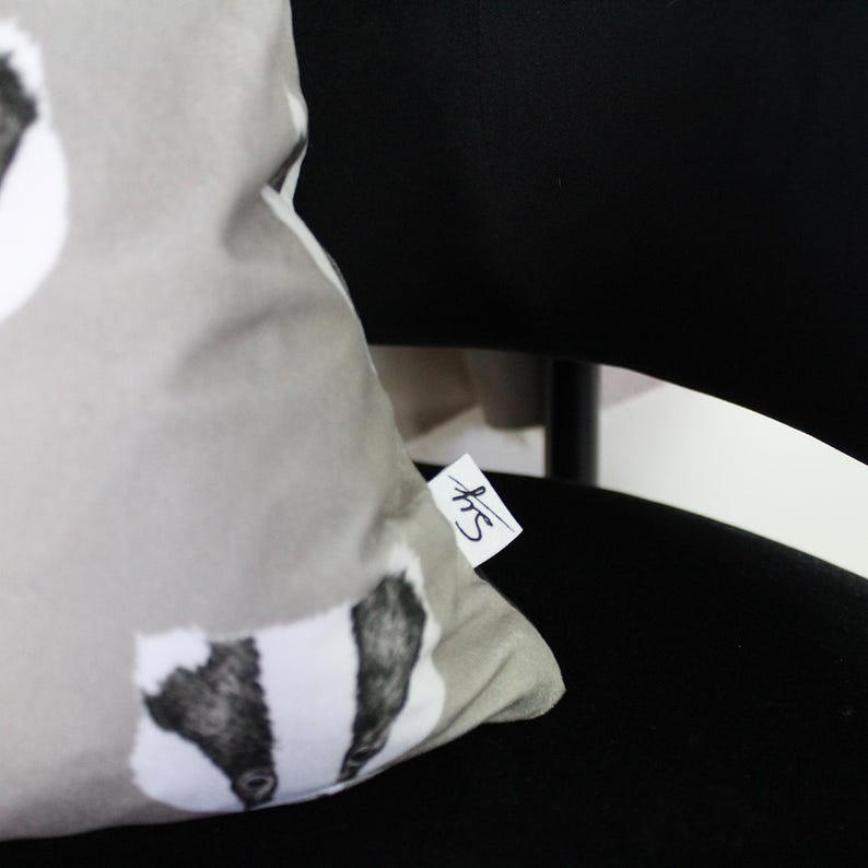Puede incluir: Un coj&iacute;n decorativo gris claro con un dise&ntilde;o de tej&oacute;n. El tej&oacute;n tiene marcas en blanco y negro. El coj&iacute;n est&aacute; sobre una silla negra. Se adjunta una peque&ntilde;a etiqueta con texto manuscrito.