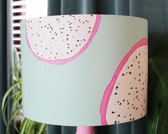 May include: A cylindrical lampshade featuring a light mint green background with a repeating pattern of sliced dragon fruit. The fruit slices have pale pink flesh, dark pink outer edges, and small black seeds scattered throughout the surface.