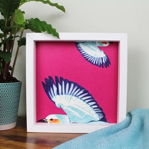 Peut inclure: Une image encadr&eacute;e blanche avec un fond rose et deux oiseaux bleus et blancs en vol. Les oiseaux sont stylis&eacute;s et ont un design graphique.