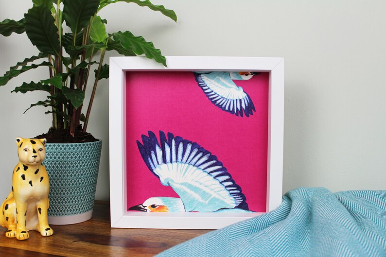Peut inclure: Une image encadr&eacute;e blanche avec un fond rose et deux oiseaux bleus et blancs en vol. Les oiseaux sont stylis&eacute;s et ont un design graphique.