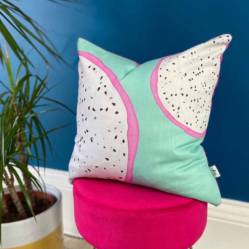 May include: A green and pink cushion with a white dragon fruit design. The cushion is sitting on a pink velvet stool.