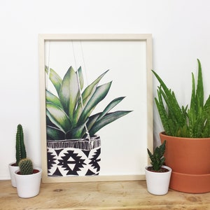 Puede incluir: Una impresi&oacute;n enmarcada de una planta verde en una maceta colgante geom&eacute;trica en blanco y negro. La impresi&oacute;n est&aacute; sobre un fondo blanco y est&aacute; enmarcada en un marco de madera claro. Hay tres peque&ntilde;as plantas en maceta en un estante de madera debajo de la impresi&oacute;n.