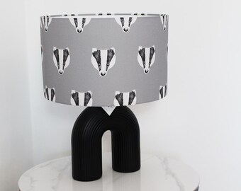 May include: A table lamp featuring a cylindrical grey lampshade with a repeating black and white badger face pattern. The lamp base is a solid black, arched, ribbed structure that provides a stable foundation for the patterned shade above.