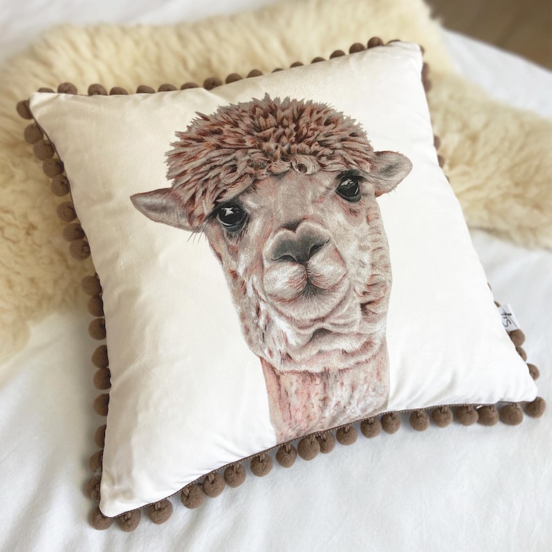 Op de afbeelding: Een wit kussen met een bruine pomponrand heeft een close-up illustratie van een bruine alpaca met een pluche kophaar.
