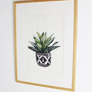 Op de afbeelding: Een ingelijste print van een aquarelillustratie van een groene plant in een zwarte en witte gepatroneerde hangende pot.