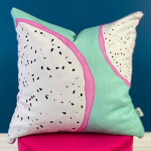 May include: A green and white cushion with a pink outline features a dragon fruit design. The cushion is sitting on a bright pink surface.