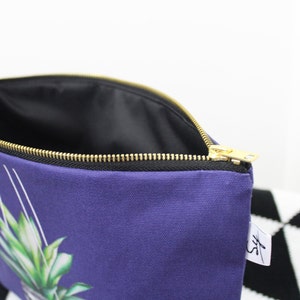 Op de afbeelding: Een paarse ritszak met een ananasprint en een zwarte rits. De zak heeft een zwarte voering en een wit label met de letters "SF".