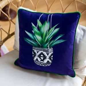 Op de afbeelding: Een blauw fluwelen kussen met een groene rand heeft een afbeelding van een hangende plant met groene bladeren in een zwart-wit gepatroneerde pot.
