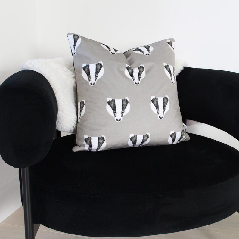 Puede incluir: Un coj&iacute;n decorativo cuadrado con un patr&oacute;n repetido de caras de tej&oacute;n en blanco y negro sobre un fondo gris. El coj&iacute;n est&aacute; colocado sobre un sill&oacute;n de terciopelo negro, con un coj&iacute;n blanco y esponjoso visible.