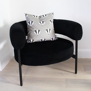 Puede incluir: Un sill&oacute;n de terciopelo negro con asiento redondo y brazos curvos. Un coj&iacute;n gris con un patr&oacute;n repetido de caras de tej&oacute;n descansa sobre la silla. La silla tiene patas de metal negro.
