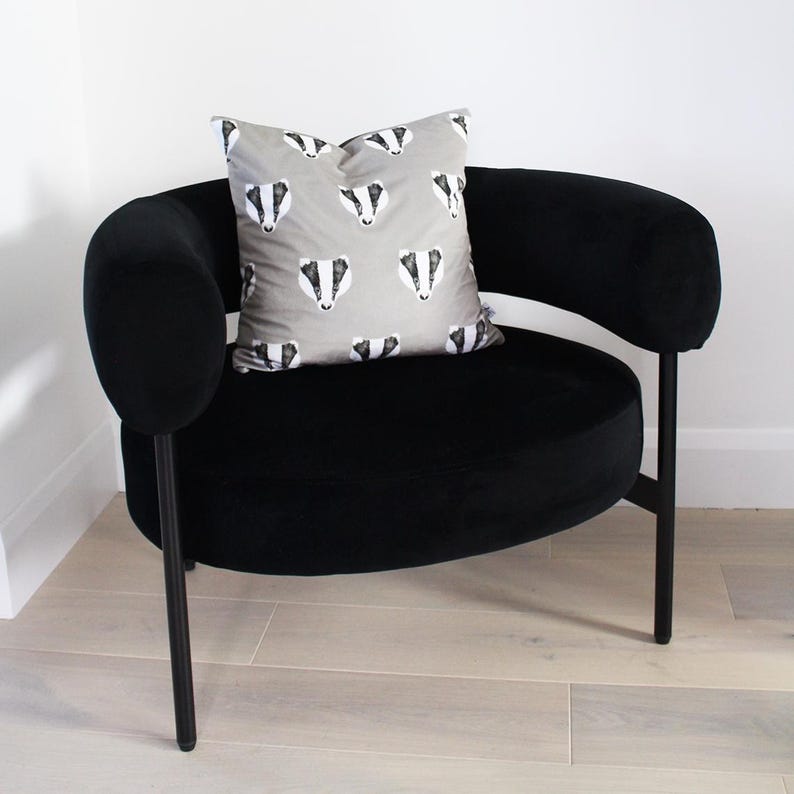 Puede incluir: Un sill&oacute;n de terciopelo negro con asiento redondo y brazos curvos. Un coj&iacute;n gris con un patr&oacute;n repetido de caras de tej&oacute;n descansa sobre la silla. La silla tiene patas de metal negro.