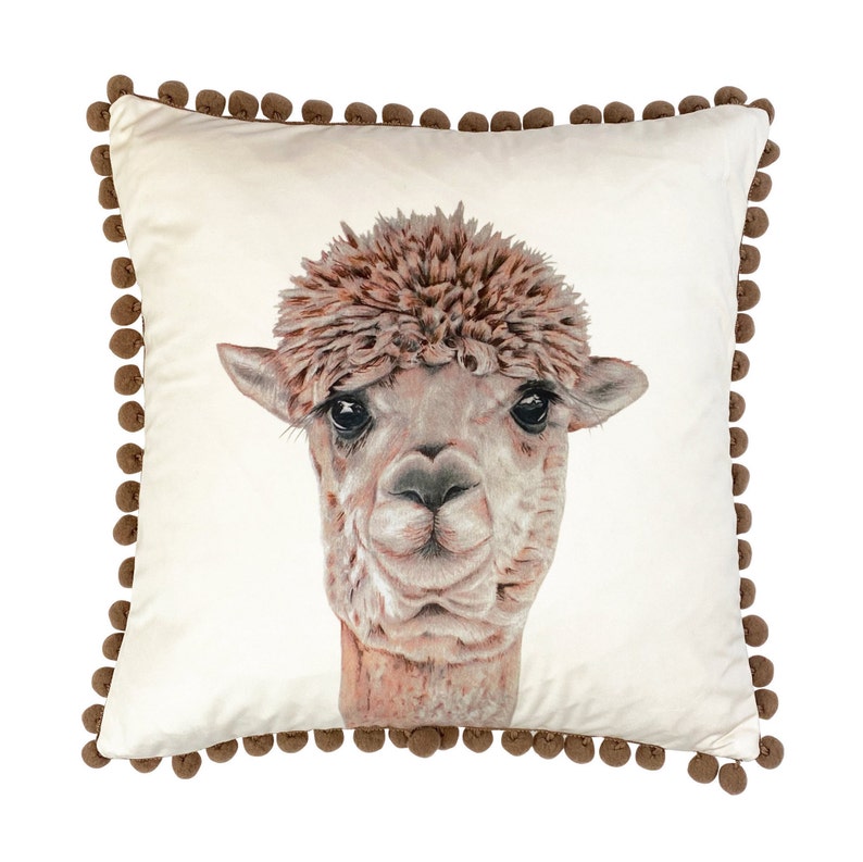 Op de afbeelding: Een wit kussen met een bruine pomponrand heeft een close-up illustratie van het gezicht van een alpaca. De alpaca heeft bruin haar en een pluche manen.