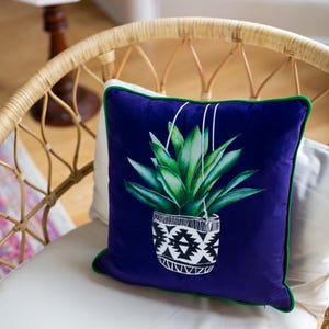 Op de afbeelding: Een kussen van paars fluweel met een groene rand heeft een grafische print van een hangende plant in een geometrische pot in zwart en wit.