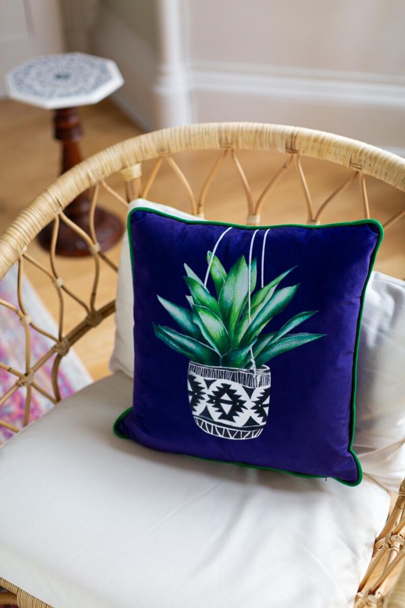 Op de afbeelding: Een kussen van paars fluweel met een groene rand heeft een grafische print van een hangende plant in een geometrische pot in zwart en wit.