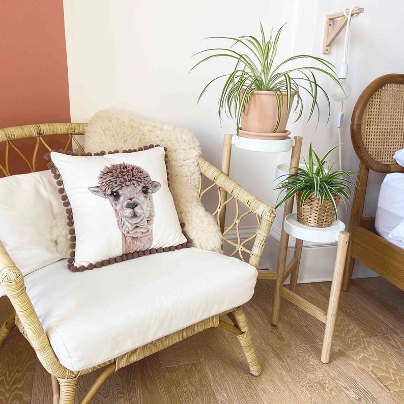 Op de afbeelding: Een witte rieten stoel met een kussen met bruine pomponrand, waarop een alpaca-illustratie is afgebeeld. De stoel staat in een kamer met een plantenstandaard waarop twee planten in potten staan, een met groene bladeren en de andere met bonte bladeren.