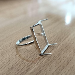 May include: Silver ring setting with a rectangular bezel for a gemstone. The ring band is plain and the bezel is open on the sides.