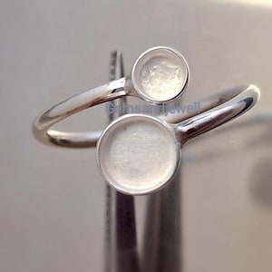 Pode incluir: Um anel prateado com duas configurações circulares. A banda do anel curva-se, com as configurações posicionadas em lados opostos. As configurações parecem ser projetadas para conter pequenas pedras preciosas redondas ou outros elementos decorativos. O anel é mostrado contra um fundo desfocado.